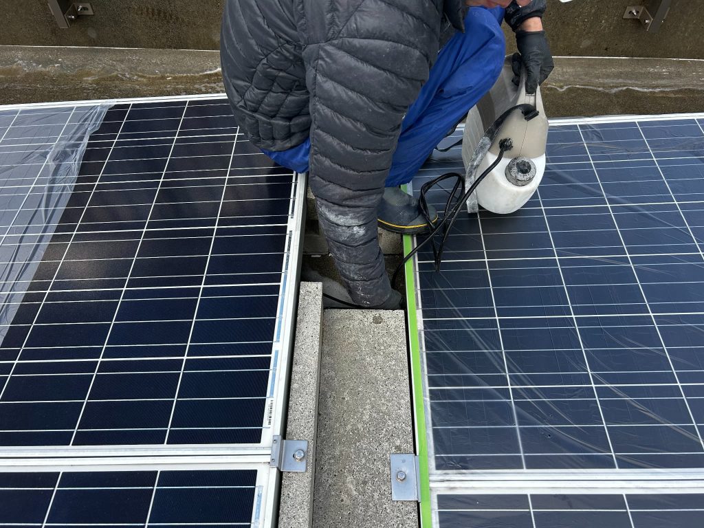 太陽光パネルの下に長ノズルを使い、シリカテックを散布してコンクリート床面を防水施工する工程